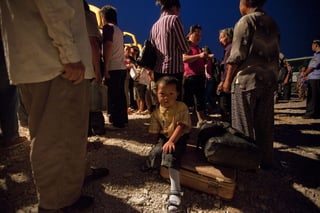 Migración. Un niño migrante permanece sentado sobre una maleta mientras aldeanos se reúnen al amanecer para continuar.