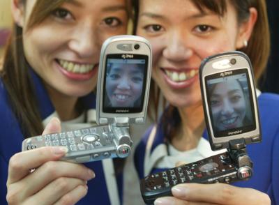 Dos jóvenes japonesas muestran sus fotografías tomadas con el nuevo teléfono celular 3G modelo P2102v. Las fotografías tomadas con este celular pueden ser enviadas por e-mail aún cuando se está hablando con otra persona por teléfono. Con este modelo también es posible grabar video. No ha sido establecido el precio de este artículo pero estará a la venta en el mercado japonés en Enero del 2003