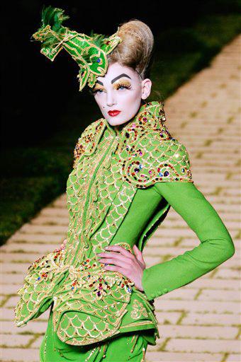 Mujeres con trompetas medievales adornando el tocado de una falda verde manzana Alicia de las Maravillas con voluminosos tops y boleros damero, amarillo y lila, rosa y amarillo o turquesa y amarillo, a juego con medias y zapatos puntiagudos, cada uno de un color, con tacones de cristal transparente