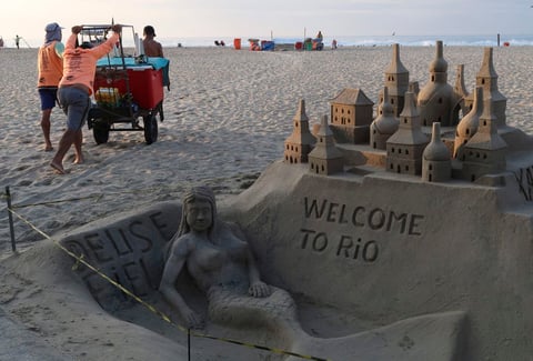 Hombres brasileños empujan su carro con refrescos y asientos a través de la arena cerca de una escultura de arena que da la bienvenida a la gente a Río con una sirena de bienvenida.