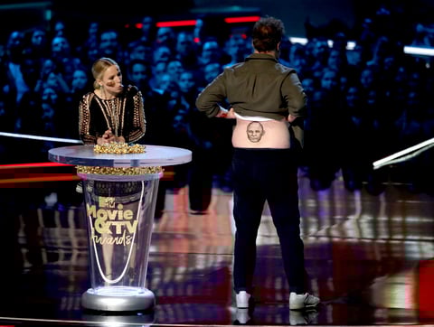 Kristen Bell, a la izquierda, observa mientras Seth Rogen muestra un tatuaje falso de Vin Diesel mientras presenta el mejor espectáculo de comedia.