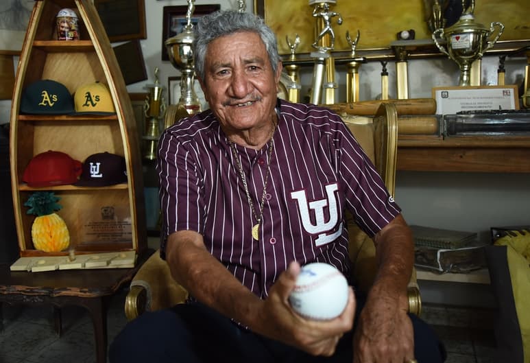 En un cuarto repleto de trofeos, Horacio Piña García sujeta la pelota para lanzar una recta.
