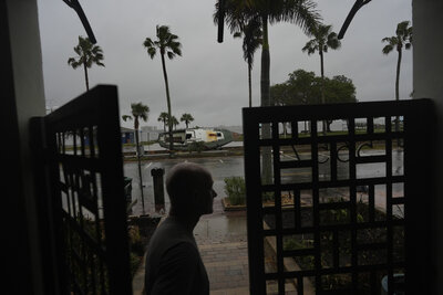 Christian Burke stands at the door of his home, where he, his mother, and his aunt plan to ride out Hurricane Milton on the third floor overlooking overlooking Tampa Bay, in Gulfport, Fla., Wednesday, Oct. 9, 2024. Burke, who said his engineer father built the concrete home to withstand a Category 5 hurricane, expects his raised ground floor to get up to 8 feet of water in Milton. A boat deposited by Hurricane Helene sits lodged in the bay front park outside his front door. (AP Photo/Rebecca Blackwell)