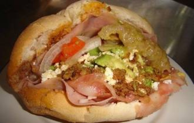 La 'Torta de la Barda' se prepara de bolillo, frijol, carne, jamón, queso amarillo, aguacate, cebolla, tomate, chorizo y chicharrón en salsa verde.